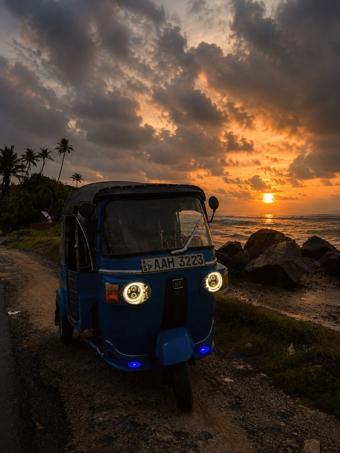 A blue tuk-tuk parked on a Weligama beach road at sunset, headlights glowing over the south-coast surf